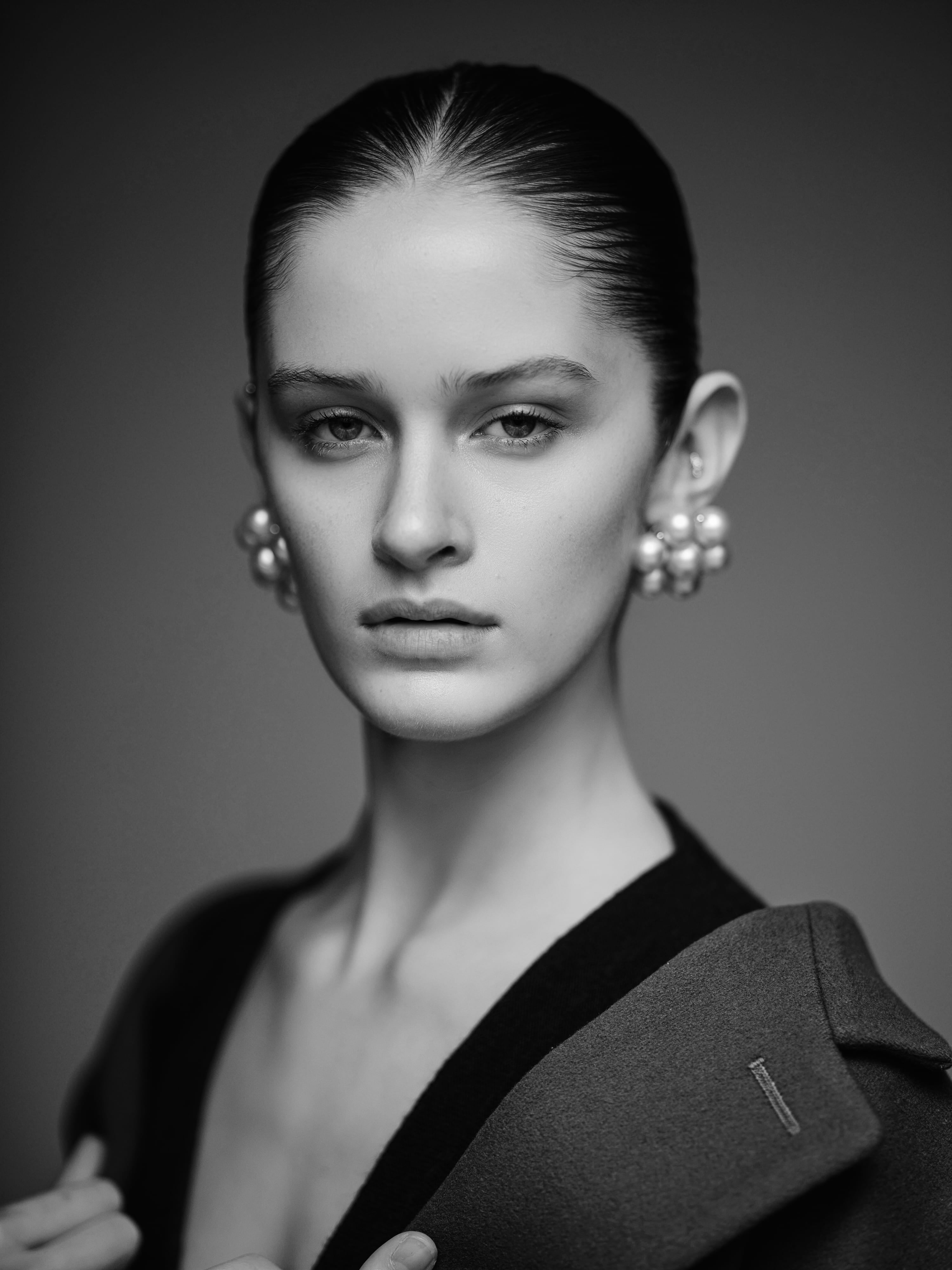 Eva with slicked-back hair and pearl cluster earrings, an oversized coat at her collar, close, black and white