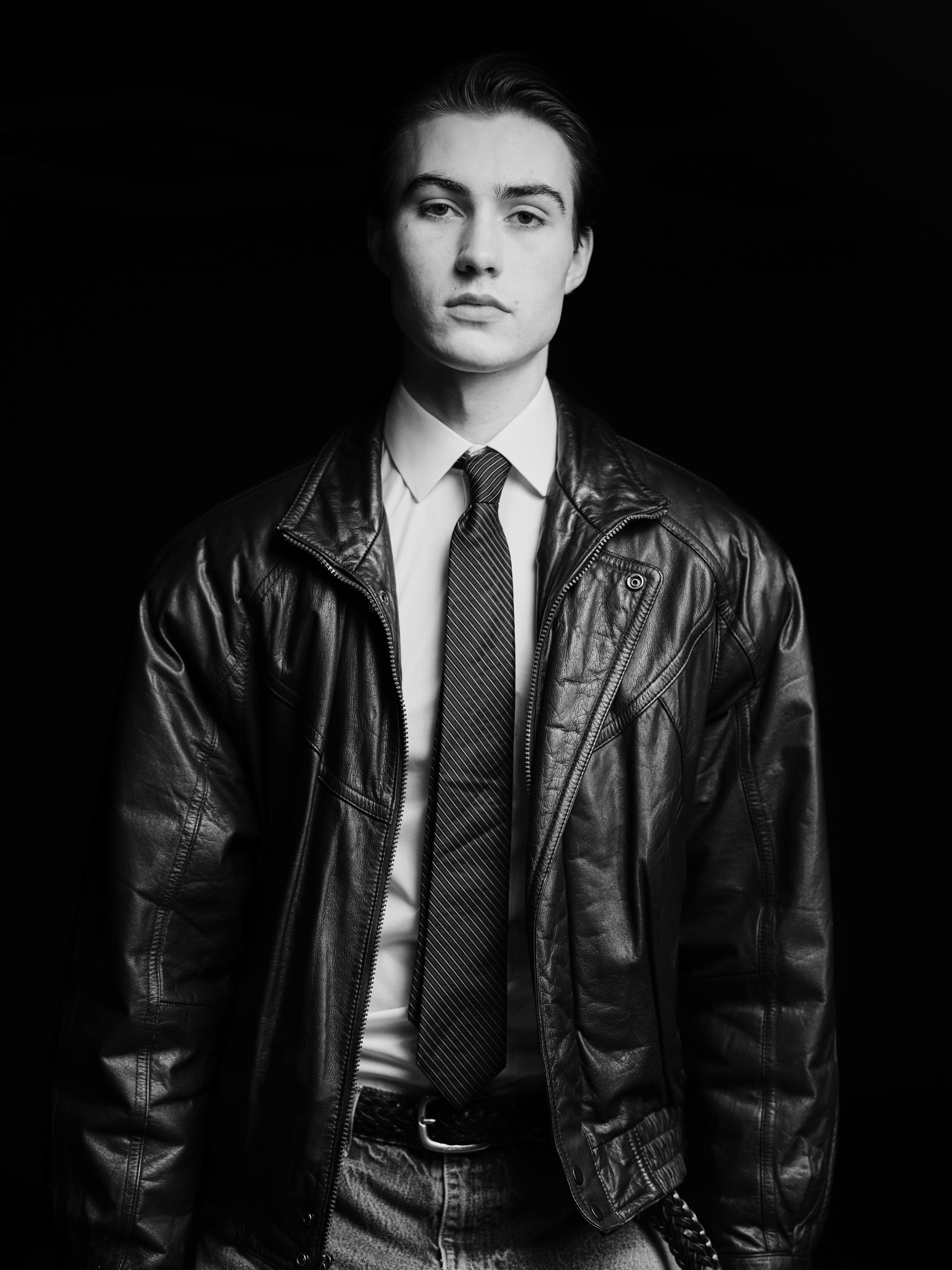 Greyson facing the camera in a leather jacket over a shirt and tie, dark ground, black and white