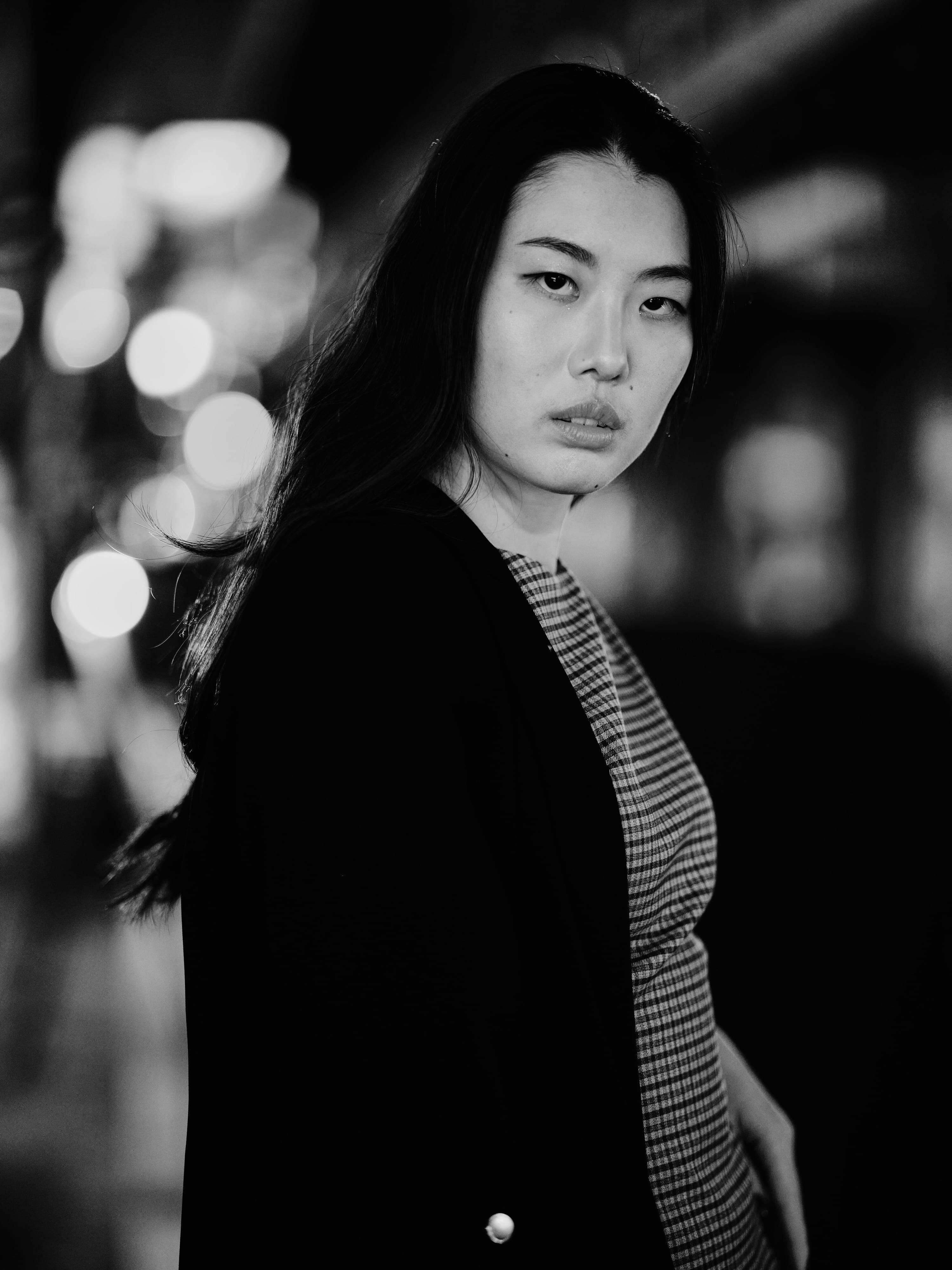 Hinano looking back over her shoulder into the camera in a black blazer, city lights blurred behind her, black and white