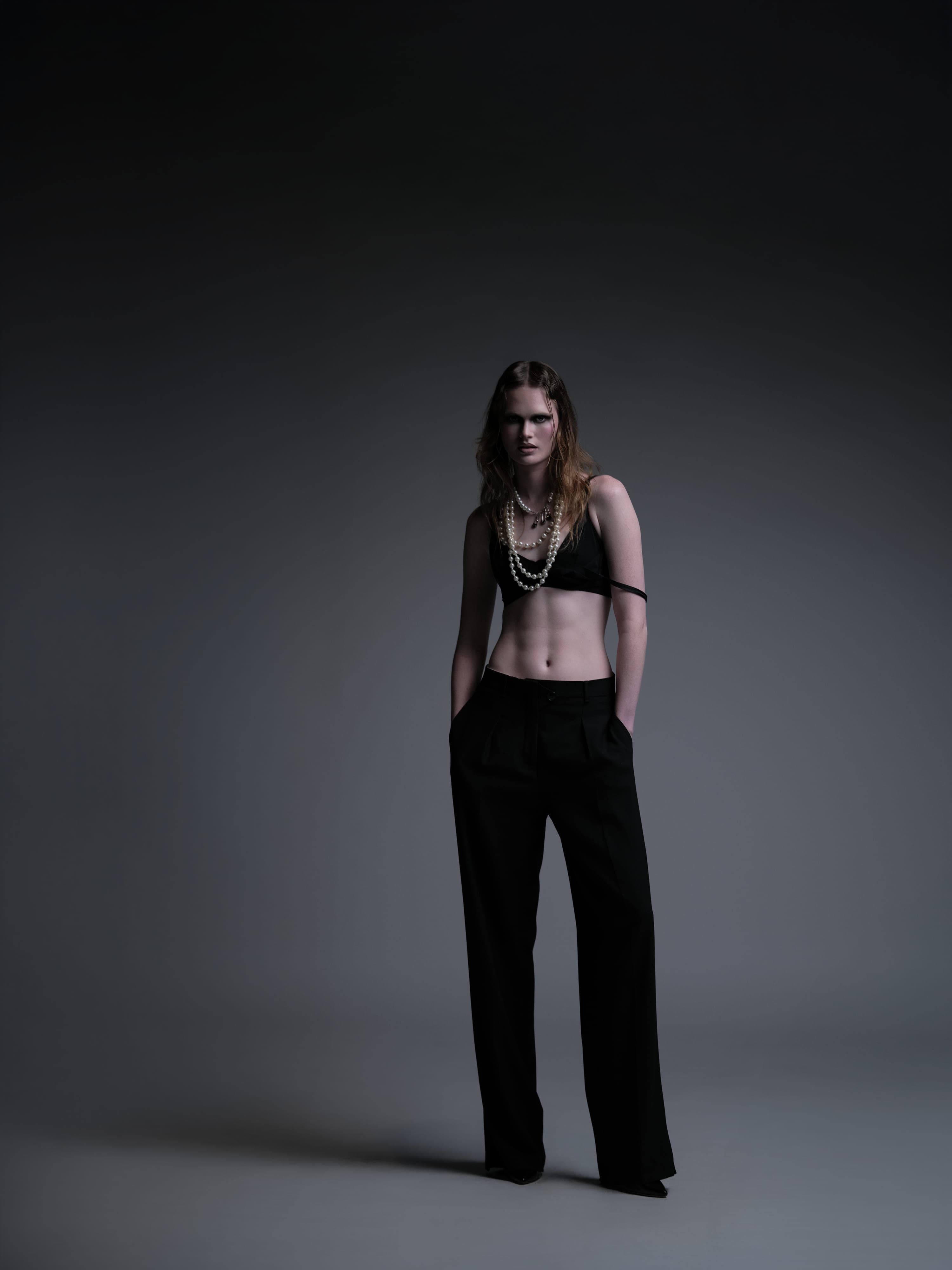 Kate Wiggins full-length studio editorial, pearl necklaces over black crop top and wide-leg trousers, dramatic overhead light