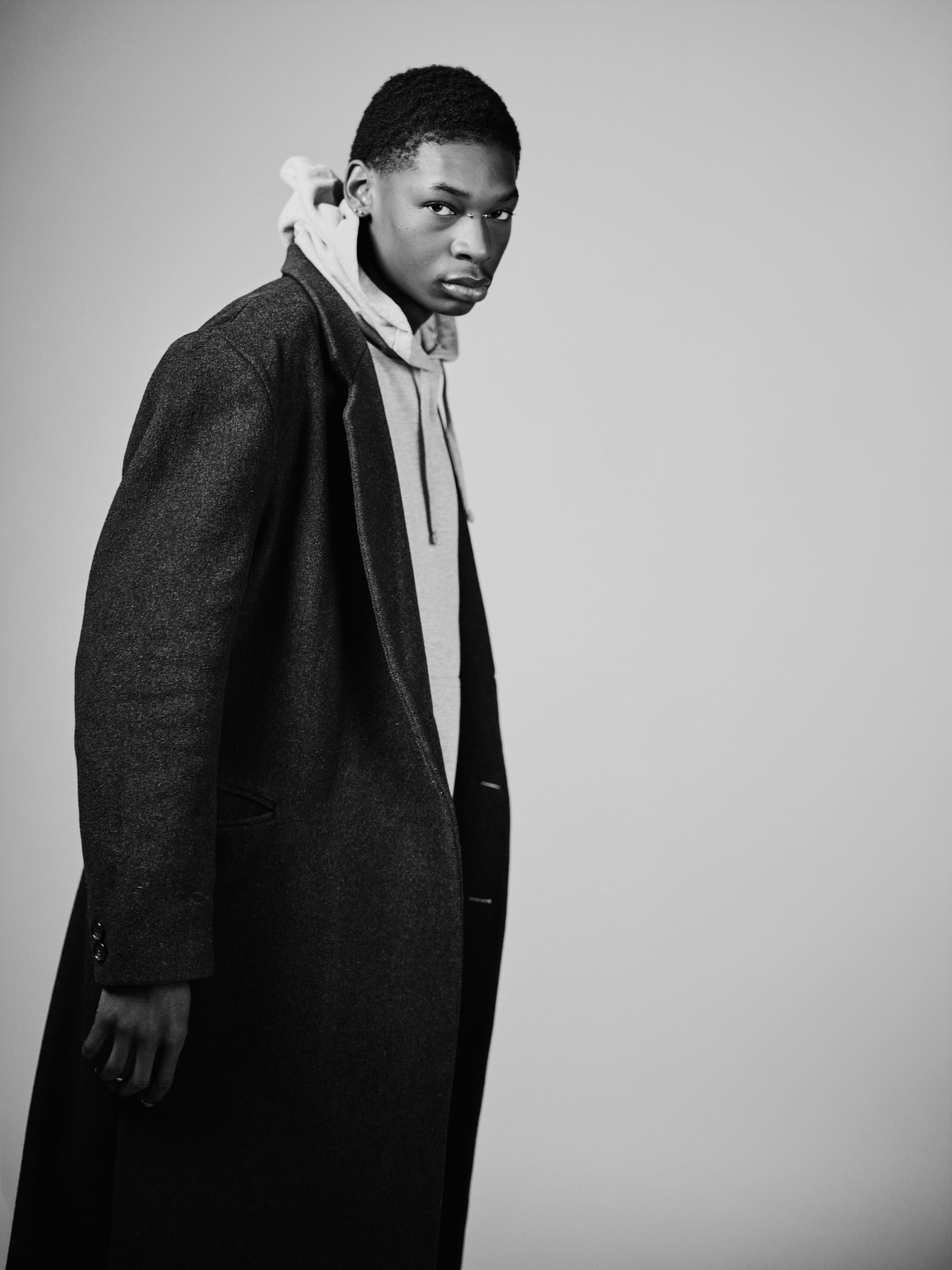 Katron in a long overcoat over a hoodie, turned three-quarters to the camera, black and white