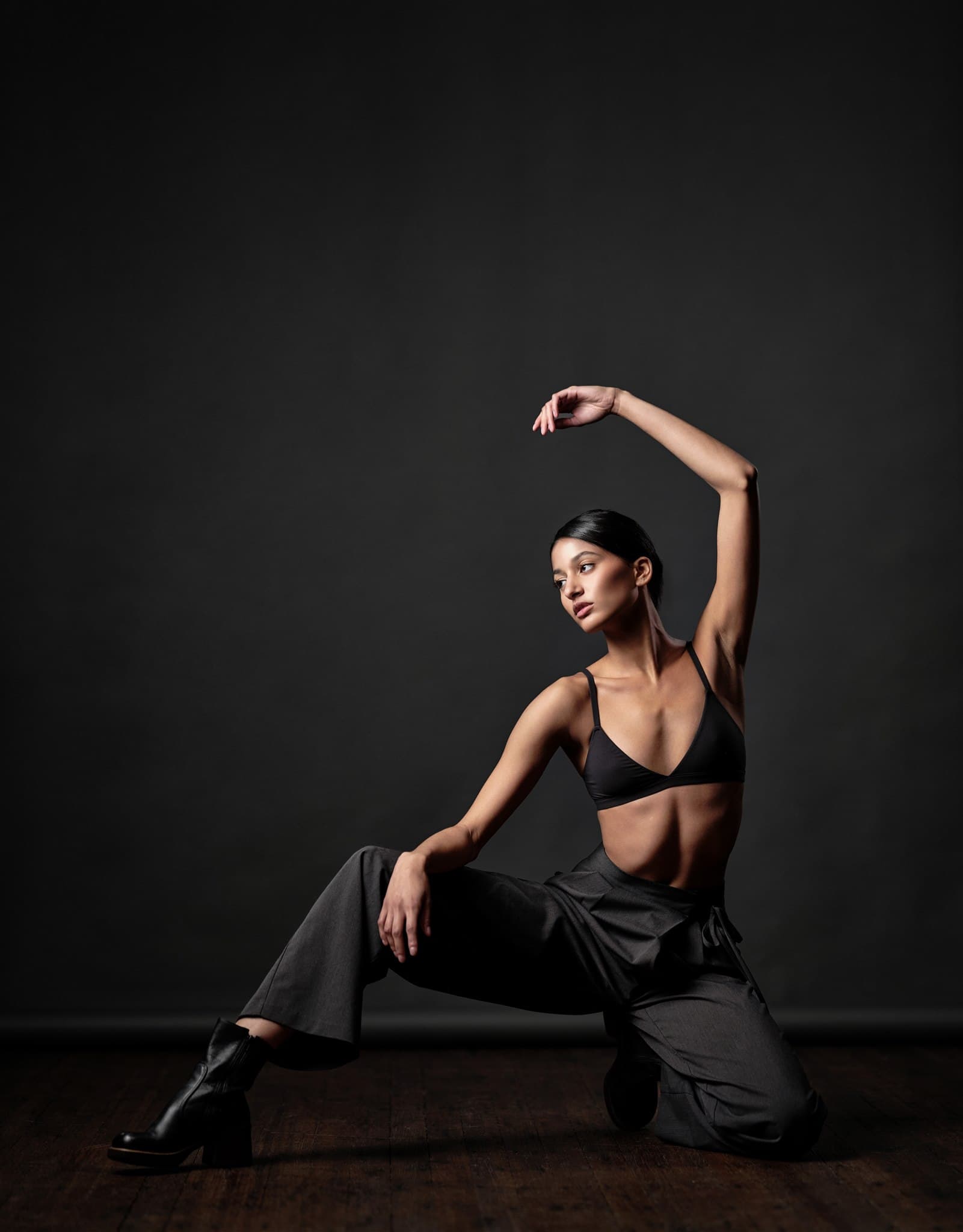 Linzy in dramatic floor pose with arm raised, black bralette and charcoal trousers, chiaroscuro studio lighting
