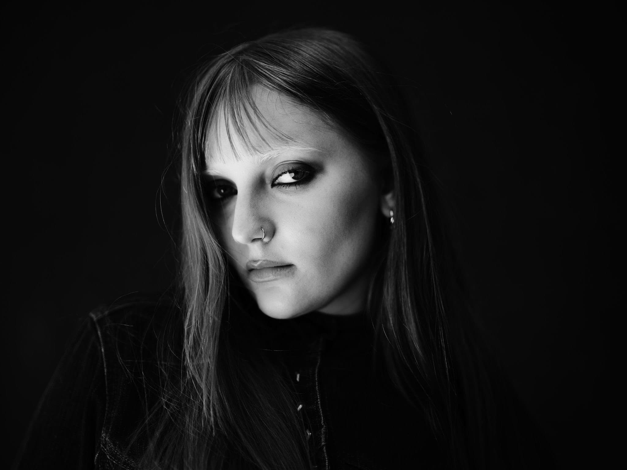 Dominique Reinhart close-up portrait, side light through bangs, dark moody background, black and white fashion editorial